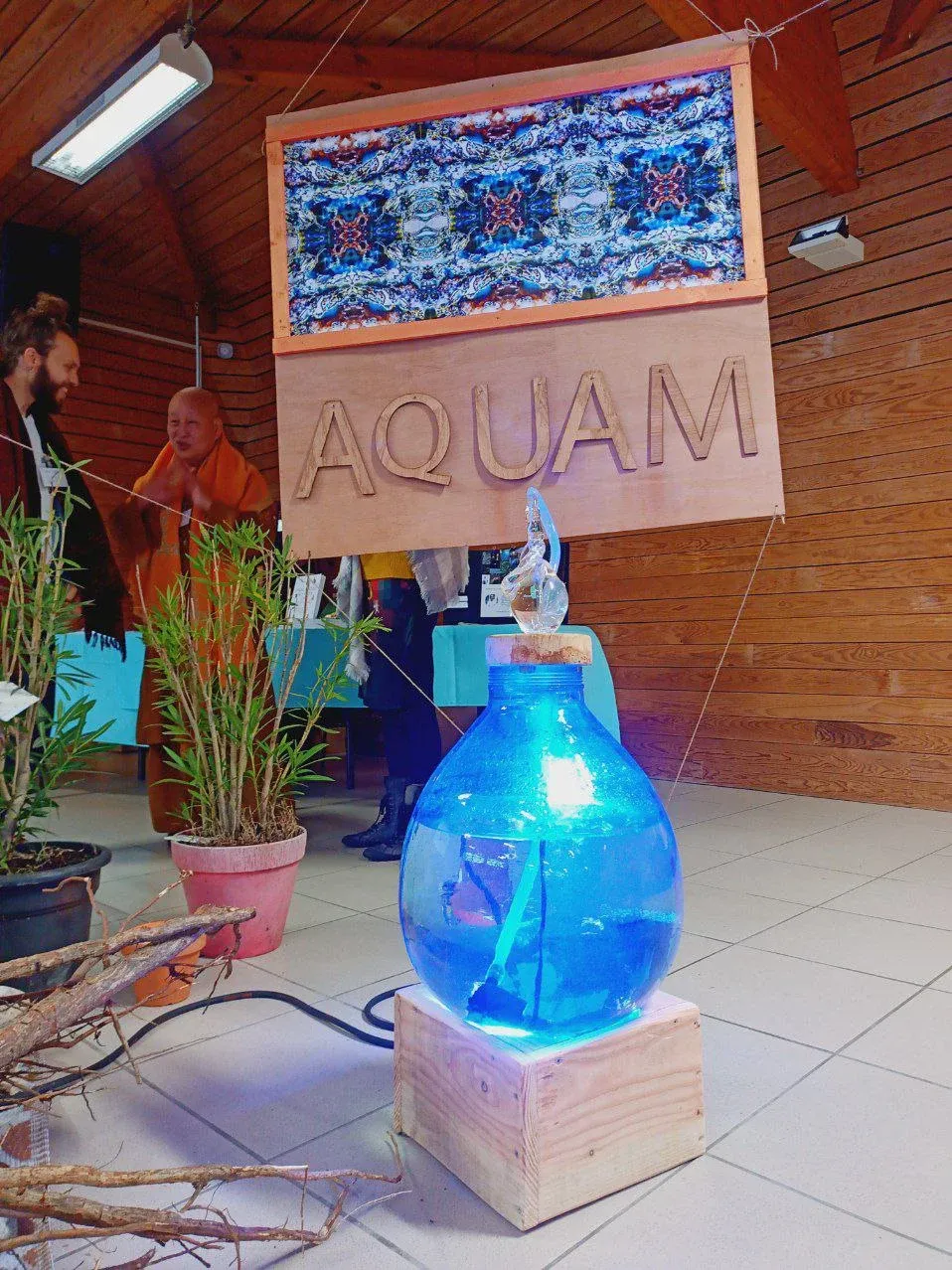 Une grande jarre de verre emplie d’eau, éclairée en bleu, surmontée d’un œuf de verre ; au fond, un moine en robe safran, mains jointes.