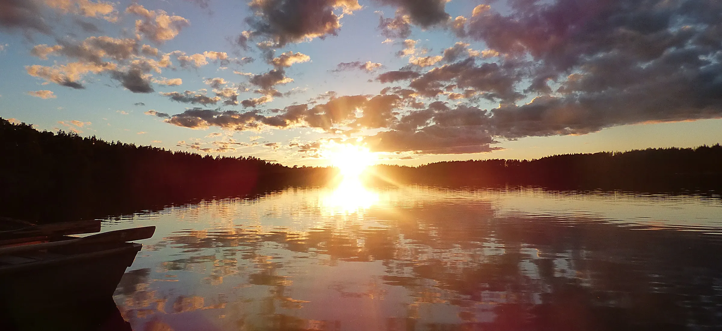 Un lac immobile au coucher du soleil, le ciel nuageux et le soleil reflétés en entier dans l’eau, l’avant d’une barque au bord de l’image.